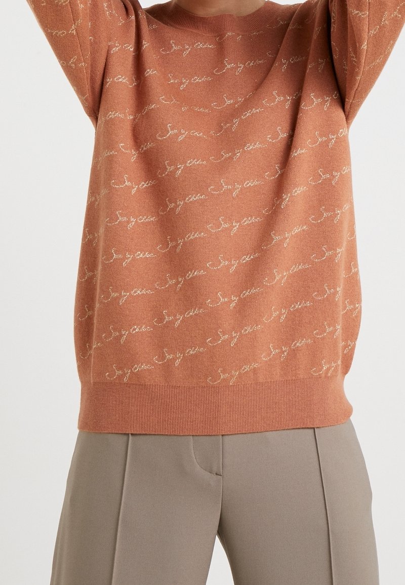Pull rouille avec un motif de texte en écriture cursive diagonale, associé à un pantalon beige taille haute, sur une personne les bras levés.