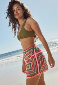 Olivenfarvet hæklede bikini top parret med en multifarvet hæklede mini nederdel, der har geometriske mønstre i rød, pink og grøn.