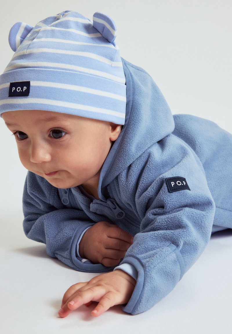 Vastasyntynyt makaamassa vatsallaan, päässään vaaleansininen raidallinen lakki korvilla ja yllään sininen fleece-takki, jossa on P.O.P.-merkit.