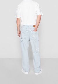 Pantalones cargo de denim azul claro con múltiples bolsillos, presentan un logo blanco en el lateral y un parche de cuero marrón en la cintura.