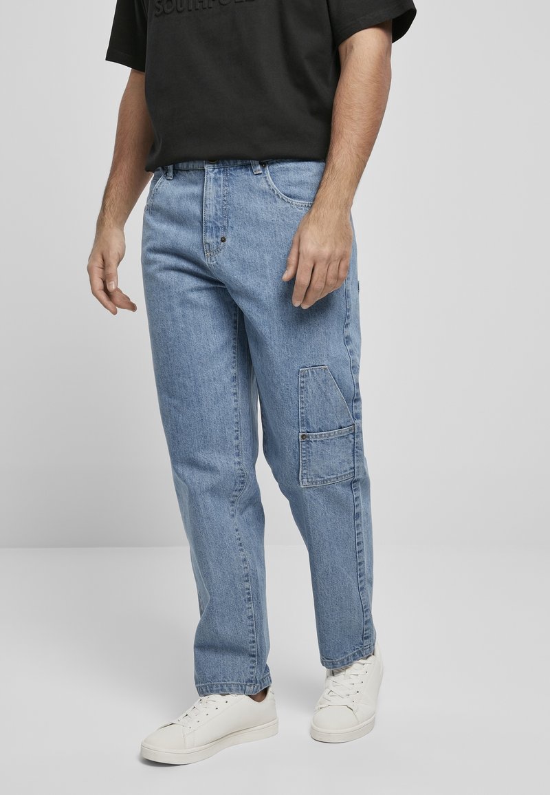 Homme portant un jean cargo en denim bleu clair avec poche latérale, un t-shirt noir à manches courtes et des baskets blanches, debout devant un fond gris.