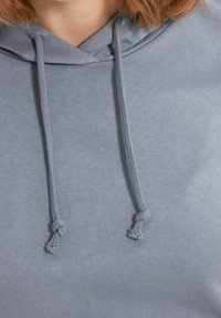 Sudadera gris claro con una textura suave, que presenta una capucha ajustable con extremos anudados. El tejido parece liso y ligeramente elástico.