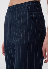 Marineblauwe broek van een gestructureerde stof, met een glinsterend streep patroon, voorplooien en zijzakken. Gesloten met een discrete rits.