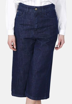 Persona che indossa jeans di denim blu scuro a gamba larga e corta e una camicia bianca a maniche lunghe con strisce verticali.