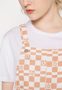 Chemise blanche sous une salopette à carreaux beige et blanc avec des motifs floraux, dotée d'un détail de bouton rond sur la bretelle.