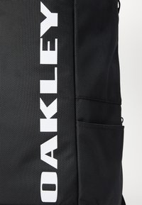 Sac à dos en tissu noir avec grand logo vertical blanc "DAKINE" et poche latérale visible à droite.