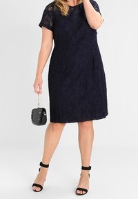 Abito in pizzo blu navy con maniche corte, linea a trapezio, motivo floreale. Borsa nera e décolleté con cavigliera completano il look.