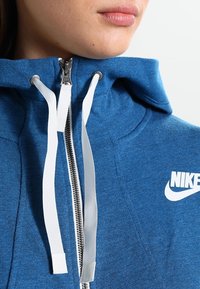 Blå zip-up huvtröja med hög krage, vitt Nike-logotyp, silverfärgad dragkedja och vita dragsnören; tyget ser mjukt ut med en melerad textur.
