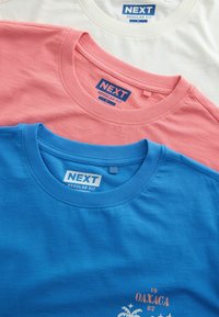 Drei gefaltete T-Shirts mit Rundhalsausschnitt in Weiß, Pink und Blau, jeweils mit "NEXT Regular Fit" in Größe Medium gekennzeichnet; das blaue Shirt hat einen teilweisen orangen Aufdruck.