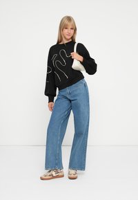 Sweatshirt noir avec des motifs de lignes décoratifs, large jean en denim bleu, et baskets beiges avec un imprimé léopard. Tient un sac à main blanc.