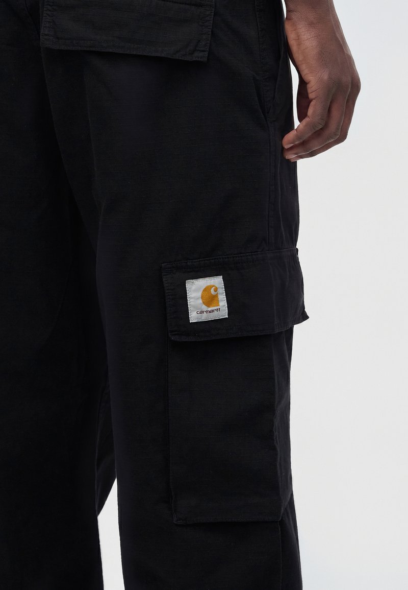 Gros plan sur la poche d'un pantalon cargo noir Carhartt avec un patch logo et la main droite d'une personne reposant à côté.