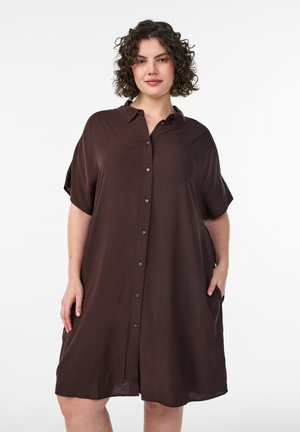 Frau mit kurzen lockigen Haaren, die ein locker sitzendes, knielanges, dunkelbraunes Hemdkleid mit kurzen Ärmeln und Kragen trägt, vor einem weißen Hintergrund stehend.