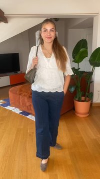 Biała bawełniana bluzka z koronkowym wykończeniem i falbaną u dołu, w zestawieniu z ciemnoniebieskimi szerokimi spodniami i szarymi butami płaskimi. Mała szara torebka i rośliny doniczkowe w tle.