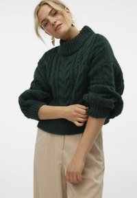 Groene gebreide trui met een hoge kraag, kabelpatroon en volumineuze mouwen, gecombineerd met lichtbeige broek.