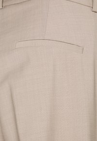 Béžové kalhoty na míru vyrobené z hladkého, strukturovaného materiálu s jemným tkaním a detailem jednoho zadního kapsy.