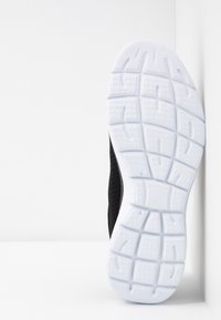 Semelle d'une chaussure de sport noir et blanc appuyée contre un mur blanc, montrant le motif texturé de la bande de roulement en dessous.