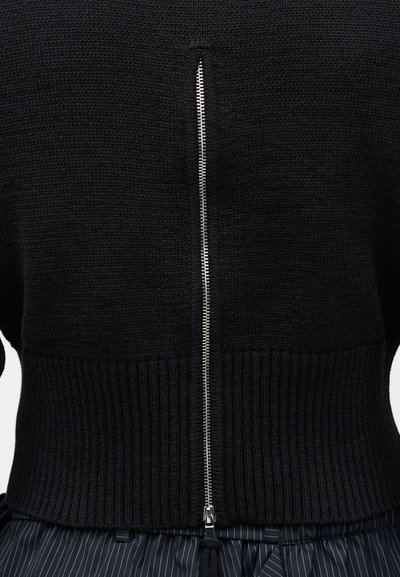 Κοντινό πλάνο μαύρου πλεκτού ενδύματος με κεντρικό ασημένιο φερμουάρ και ριμπ τελείωμα, σε συνδυασμό με σκούρο παντελόνι με λεπτές ριγές.