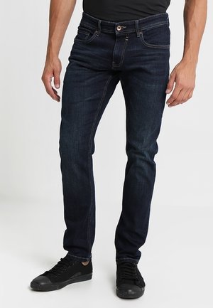 Mężczyzna w ciemnoniebieskich jeansach slim-fit i czarnych sportowych butach stoi na jasnym tle, z rękami swobodnie opuszczonymi wzdłuż ciała.