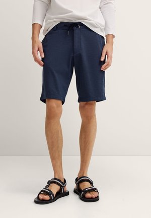 Man wearing navy blue drawstring shorts, white long-sleeve shirt, and black open-toe sandals standing on light floor.