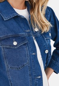 Tumma sininen farkkutakki hopeisilla napin kiinnikkeillä, klassisella kauluksella, etutaskuilla ja hieman kuluneella pinnalla.