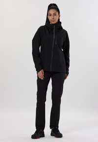 Veste noire imperméable avec un col haut et une fermeture éclair sur toute la longueur. Associée à un pantalon et des chaussures noirs. Texture lisse, design minimaliste.