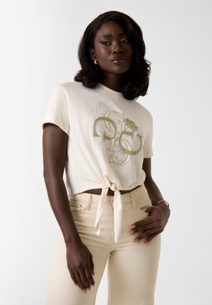 Vrouw die een beige gebonden T-shirt met print draagt en bijpassende broek met hoge taille, poserend met één hand in de zak en de andere rustend op haar dij.