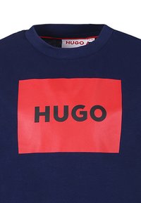 Marineblauwe katoenen t-shirt met een grote rode rechthoekige grafiek met de tekst "HUGO" in vetgedrukte zwarte lettertypes in het midden.