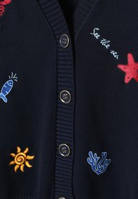 Cardigan bleu marine avec boutons cerclés d'or, présentant une broderie colorée sur le thème de la mer : poissons, soleil, corail, étoile de mer rouge et le texte « See the sea ».