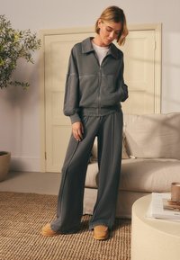 Donna in giacca zip-up grigia scura e pantaloni a gamba larga, in piedi su un tappeto intrecciato vicino a un divano beige e un tavolino rotondo con libri e una tazza.