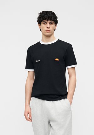 Jauns vīrietis ar cirtainiem tumšiem matiem, tērpies melnā T-kreklā ar baltu apmalējumu un pelēkās sporta biksēs, uz gaiši pelēka fona.