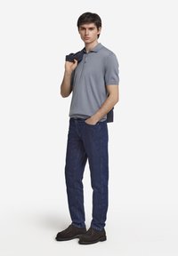 Grå polo skjorta med korta ärmar, blå denimjeans och mörkbruna skor. Modellen håller en mörk tröja hängande över ena axeln.