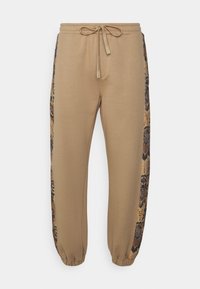 Beiga joggers med dragsko i midjan, med mönstrade sidpaneler i brunt och guld. Elastiska muddar vid anklarna förbättrar passformen.
