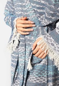 Modra in bela vzorčasta halja iz teksturiranega materiala, z detajli franja, pasom in žepom. Roke držijo tkanino halje.