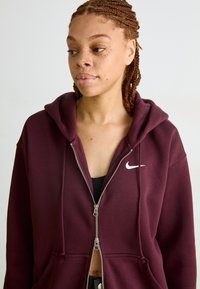 Fleece com fecho em burgundy com um logótipo Nike branco. Apresenta capuz, bolsos frontais e um tecido macio e texturizado.