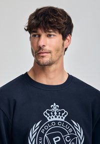 Felpa blu navy con logo ricamato bianco che presenta una corona e foglie di alloro. Collo rotondo e texture del tessuto morbido.