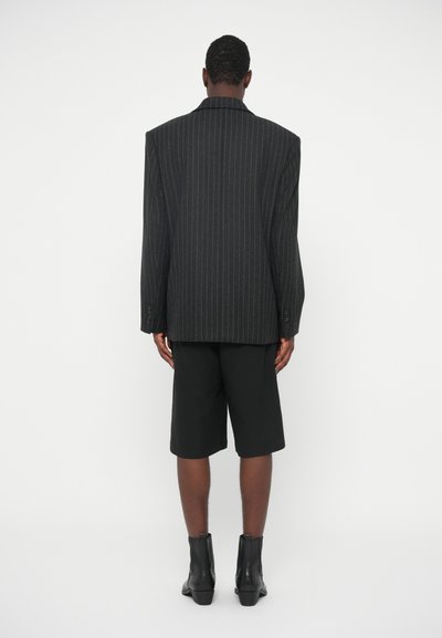 Przesadnie dużą czarną marynarkę w paski z szerokimi ramionami zestawiono z czarnymi szortami i botkami na kostkę. Gładki materiał o zdefiniowanym kształcie.