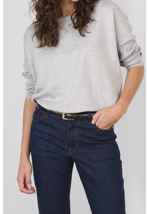 Personne portant un pull à manches longues gris clair rentré dans un jean bleu foncé avec une ceinture noire, une main dans la poche, cheveux bouclés visibles.