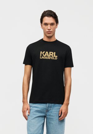 Homme portant un T-shirt noir à manches courtes avec le logo doré "KARL LAGERFELD" et un jean bleu clair, debout devant un fond clair uni.