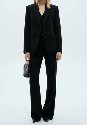 Personne portant un costume noir sur mesure avec un pantalon à jambes larges et des chaussures pointues, tenant un petit sac à main noir avec des poignées métalliques rondes.