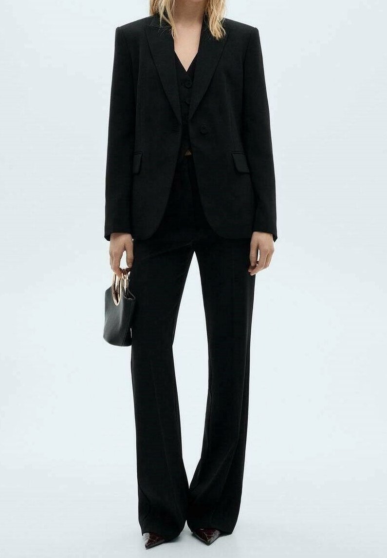 Personne portant un costume noir sur mesure avec un pantalon à jambes larges et des chaussures pointues, tenant un petit sac à main noir avec des poignées métalliques rondes.