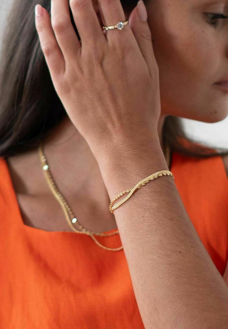 Guldarmband med en vågig design och pärlprydnader, bärs på handleden. Åtföljs av ett delikat guldhalsband. Orange topp i bakgrunden.
