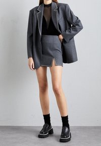 Blazer gris con detalles de tachuelas, combinado con una falda mini gris a juego con una abertura frontal. Top de malla negra y zapatos negros voluminosos.