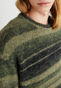 Texturerad grön stickad tröja med horisontella ränder i mörkgrön och oliv, med rund halsringning och en mjuk, fluffig yta.