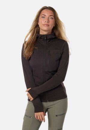 Joven mujer con cabello rubio largo lleva una chaqueta negra con cremallera y pantalones verde oliva con bolsillos con cremallera, de pie frente a un fondo liso.