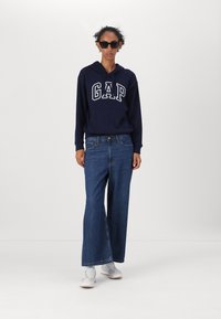 Marinblå hoodie med "GAP"-logotyp, blå vida jeans och ljusa sneakers. Modellen bär svarta solglasögon och har lockigt hår. Mjuk tygtextur.