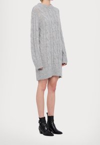 Personne portant une robe-pull oversize gris en maille torsadée, associée à des bottines noires à bout pointu et à talons.
