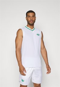 Weißes ärmelloses Basketballtrikot mit grünen Akzenten am Kragen und Logo. Kombination mit weißen Shorts, die ein grünes Logodetail aufweisen.