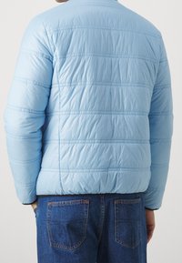 Ljust blå pufferjacka med horisontell sömnad, rund krage och elastiska ärmar. Bärs med blå jeans med synliga fickor.