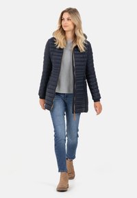 Marine blauwe pufferjack met een rits aan de voorkant, horizontaal gequilt ontwerp, en een capuchon. Draagt over een grijs shirt en blauwe jeans, gecombineerd met beige enkellaarsjes.