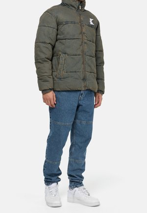 Padded jacket in olive green with a front zipper, two side pockets, and a logo patch. Paired with blue denim jeans and white sneakers.